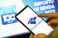 Mão usando smartphone com app da Receita Federal e telas de Imposto de Renda 2022 e 100 Anos ao fundo.