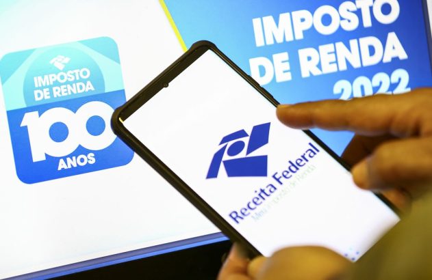 Mão usando smartphone com app da Receita Federal e telas de Imposto de Renda 2022 e 100 Anos ao fundo.