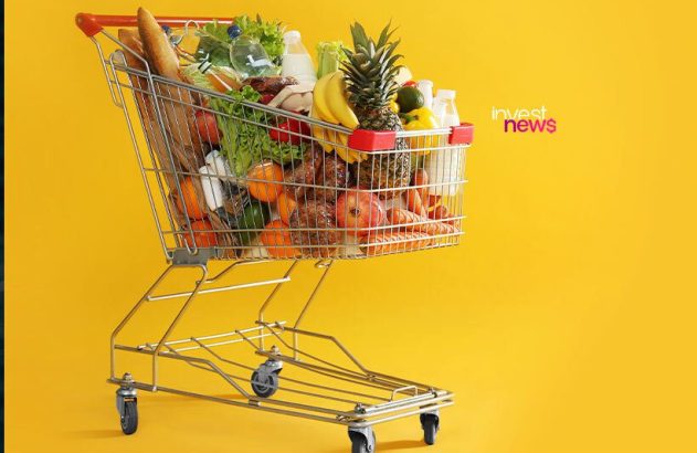 Carrinho de compras lotado com frutas, vegetais e alimentos variados em fundo amarelo.