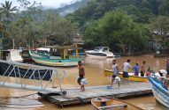 Homens carregam suprimentos em cais flutuante em rio barrento, com barcos e vegetação ao fundo.