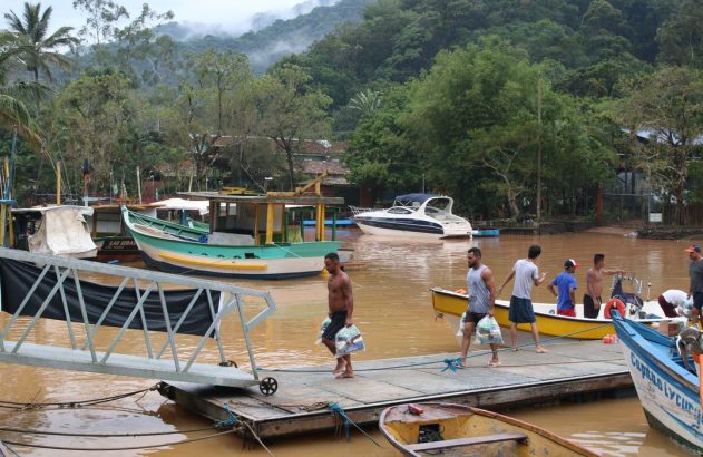 Homens carregam suprimentos em cais flutuante em rio barrento, com barcos e vegetação ao fundo.