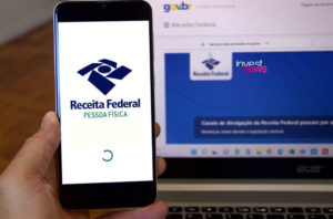 uma mão segurando um smartphone com a tela mostrando o aplicativo da Receita Federal. Ao lado, um notebook mostrando o site da Receita Federal