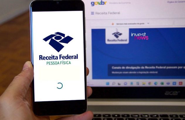 uma mão segurando um smartphone com a tela mostrando o aplicativo da Receita Federal. Ao lado, um notebook mostrando o site da Receita Federal