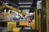 Braço robótico amarelo FANUC segura caixa em fábrica automatizada.