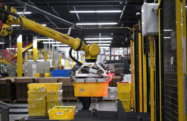 Braço robótico amarelo FANUC segura caixa em fábrica automatizada.