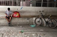 Homem sentado com mochila de entrega e bicicleta ao lado, sob estrutura urbana.