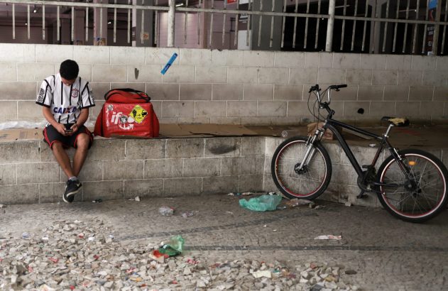 Homem sentado com mochila de entrega e bicicleta ao lado, sob estrutura urbana.