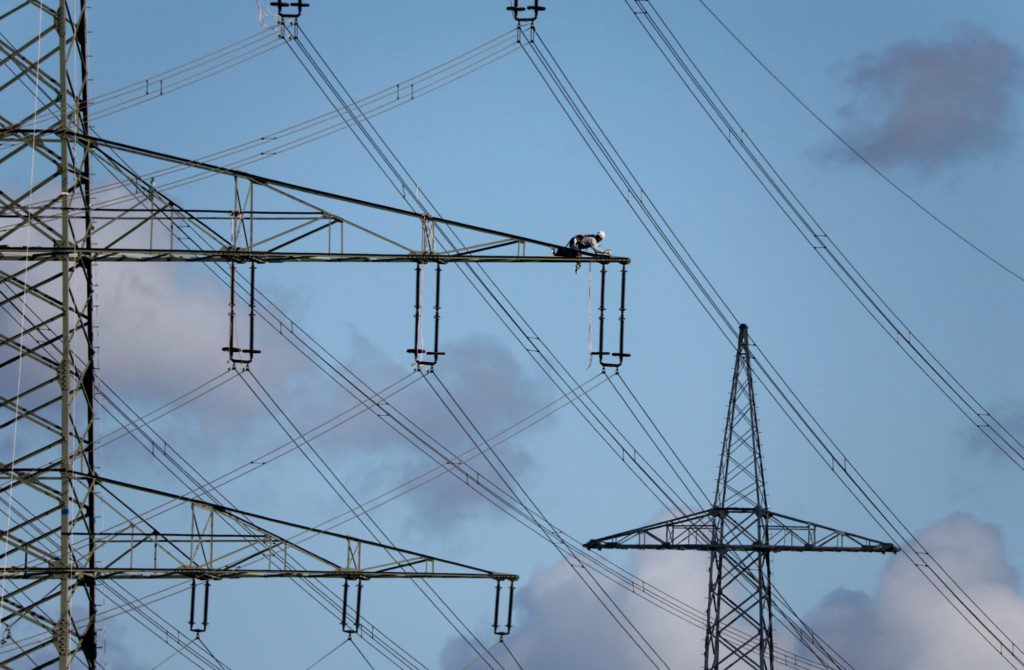 Eletricista realiza manutenção em torre de alta tensão. Fiação elétrica e outras torres sob céu azul com nuvens.