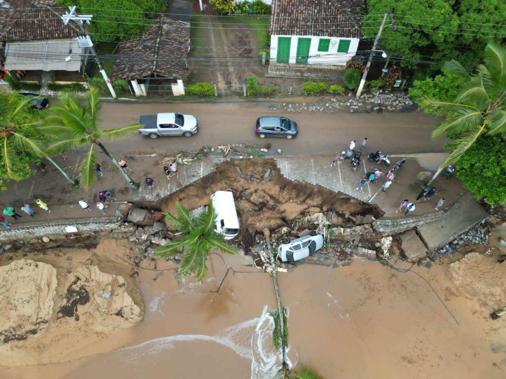 Vista aérea: estrada costeira desabou, formando cratera gigante. Carros parados no limite e um veículo caído na fenda, junto à praia.