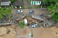 Vista aérea: estrada costeira desabou, formando cratera gigante. Carros parados no limite e um veículo caído na fenda, junto à praia.
