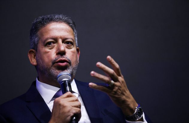 Homem de terno e barba grisalha discursa ao microfone, gesticulando. Fundo escuro.