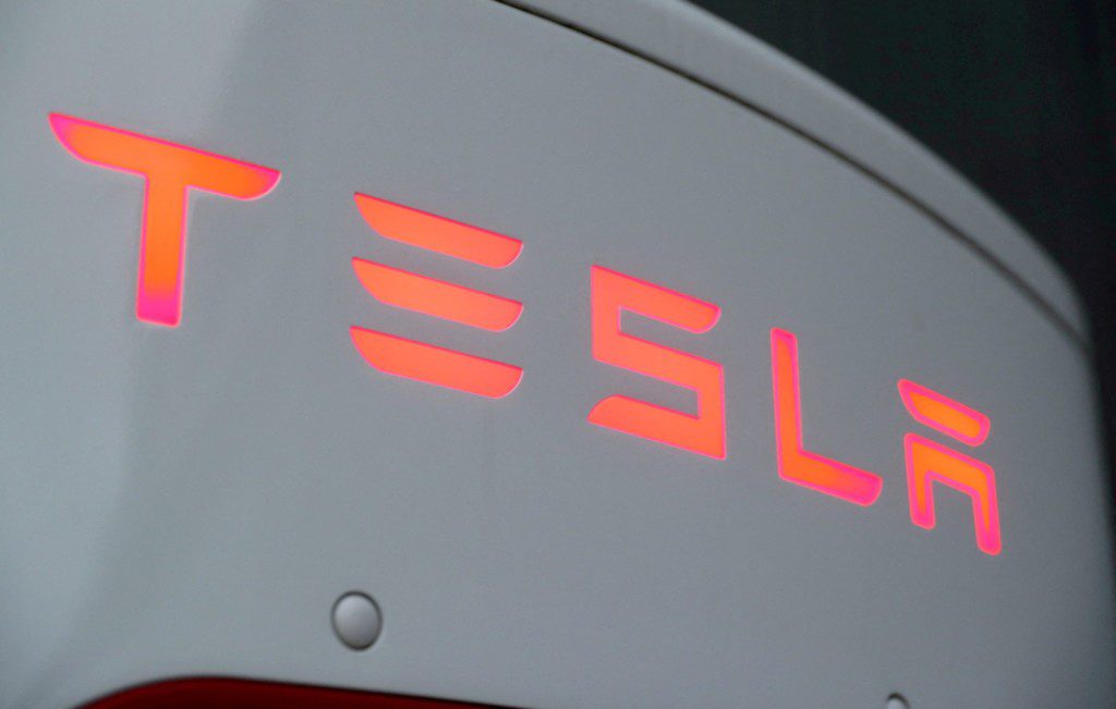 Logotipo "TESLA" iluminado em tons de laranja e rosa em carro branco.