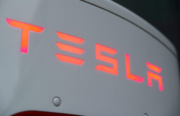 Logotipo "TESLA" iluminado em tons de laranja e rosa em carro branco.