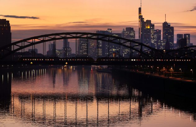 Anoitecer na cidade: ponte com trem sobre rio, reflexos na água e prédios iluminados.