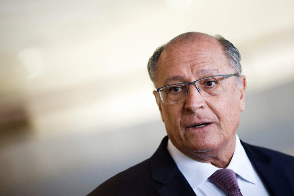 Homem de óculos, terno e gravata, com boca aberta como se estivesse falando. Fundo claro e desfocado.