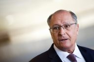 Geraldo Alckmin está de óculos, terno e gravata, com boca aberta como se estivesse falando. Fundo claro e desfocado