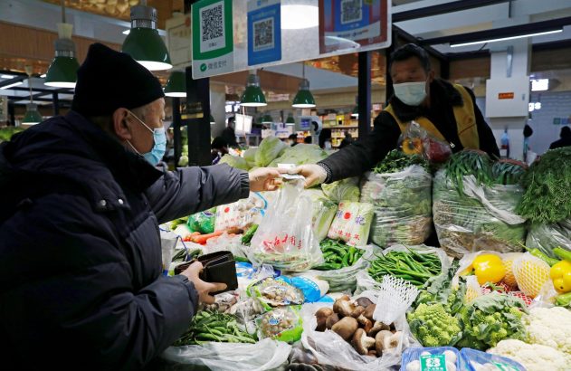 Cliente de máscara paga vendedor de máscara em feira. Balcão com variedade de legumes e verduras.