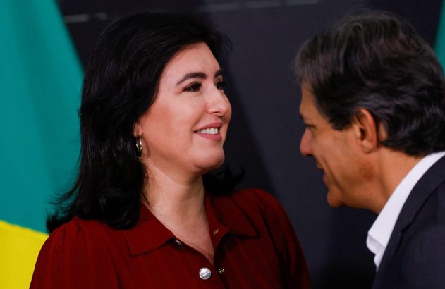 Mulher de camisa vermelha sorrindo para homem de terno em conversa. Fundo: bandeira verde e amarela.