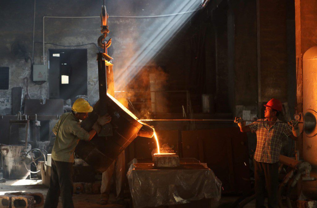 Dois trabalhadores despejam metal incandescente em uma fundição, com raios de sol e faíscas.