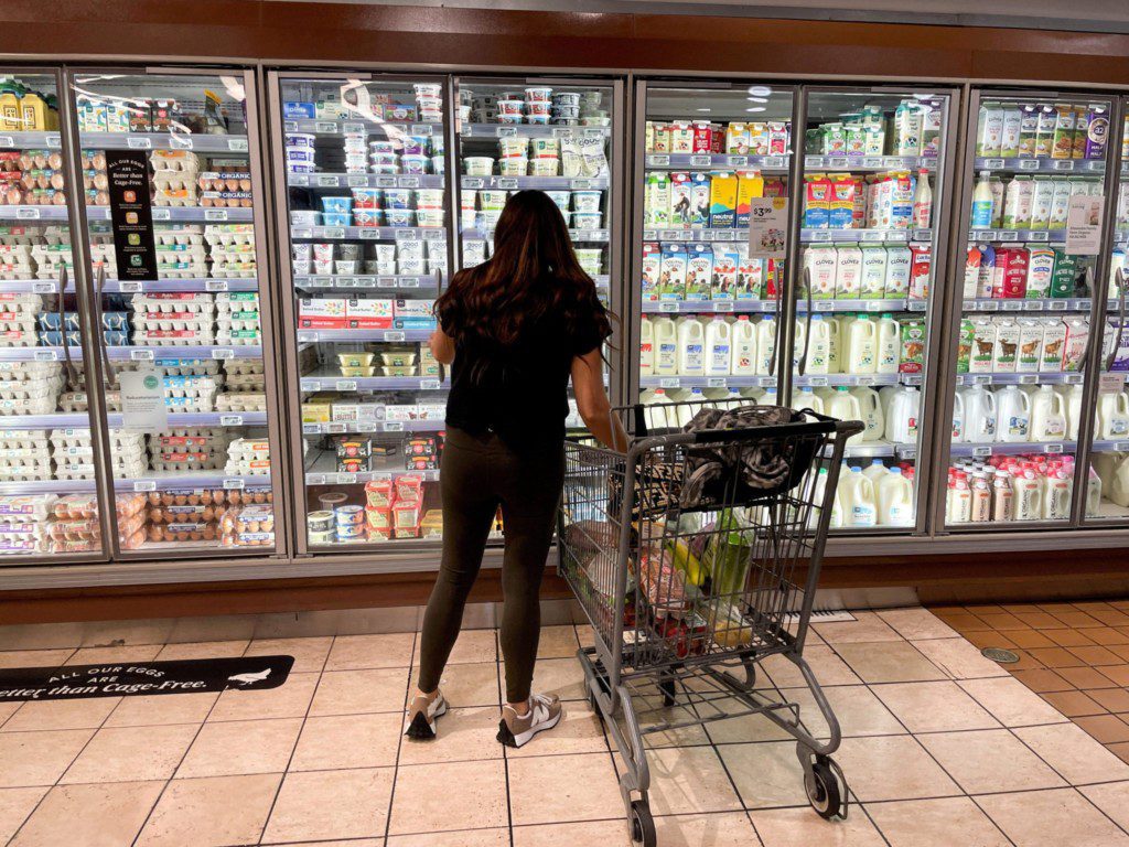 Cliente de costas em corredor de supermercado, olhando expositores de laticínios e ovos. Carrinho de compras cheio ao lado.