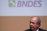 Homem de bigode e terno observando em evento do BNDES com o logo da instituição ao fundo.