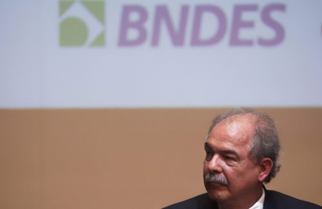 Homem de bigode e terno observando em evento do BNDES com o logo da instituição ao fundo.