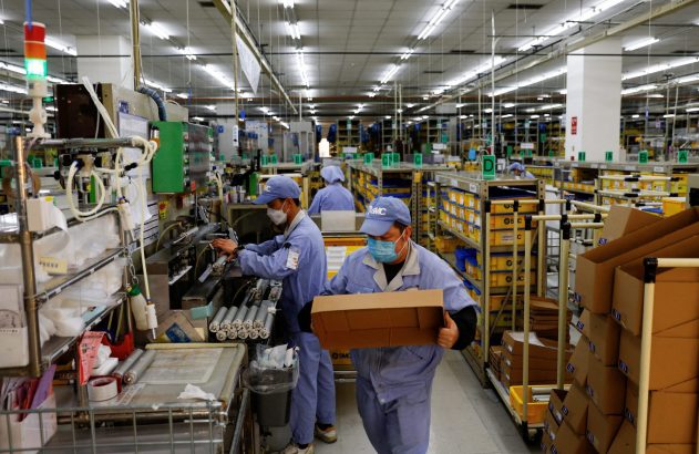 Fábrica: trabalhadores com máscaras e uniformes operam linha de montagem, um deles carrega caixa.