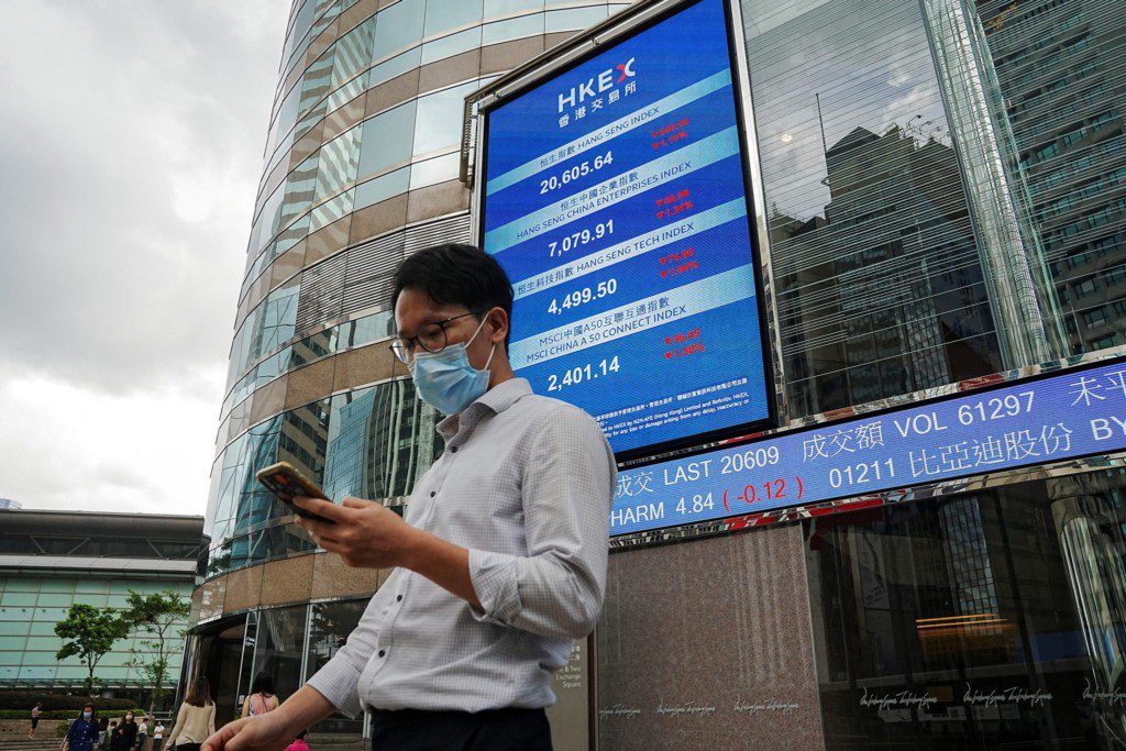 Homem de máscara e óculos checa celular; tela da bolsa de valores de Hong Kong e prédio de vidro ao fundo.