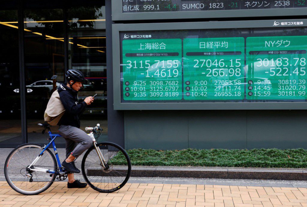 Ciclista olhando celular em frente a um painel digital exibindo cotações de bolsas de valores em queda.