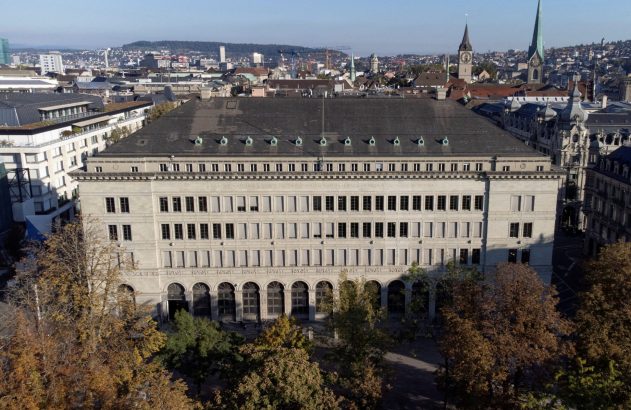 Vista aérea do Banco Nacional Suíço em Zurique, com a cidade e colinas ao fundo e árvores de outono em primeiro plano.