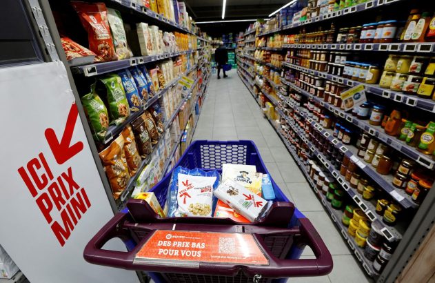 Carrinho de compras em corredor de supermercado, com prateleiras cheias de produtos e sinais de 'preços baixos'.