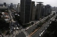 Vista aérea de avenida urbana com corredor de ônibus, tráfego intenso de veículos e pessoas, entre prédios altos.