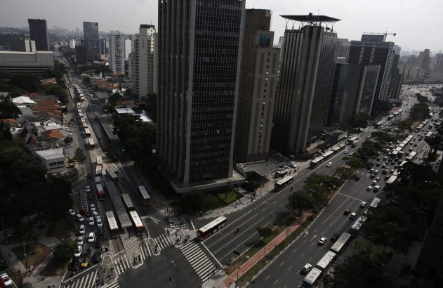 Vista aérea de avenida urbana com corredor de ônibus, tráfego intenso de veículos e pessoas, entre prédios altos.