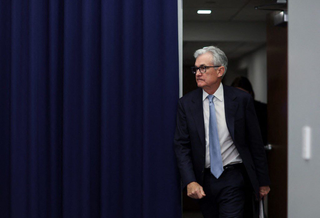 Jerome Powell em terno escuro, caminhando por porta, olhando para o lado.