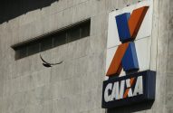 Pássaro voa perto de prédio cinza com letreiro azul e laranja da CAIXA.