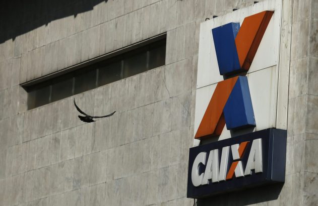 Pássaro voa perto de prédio cinza com letreiro azul e laranja da CAIXA.