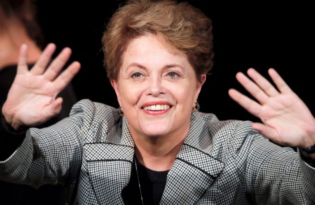 Mulher de cabelo curto e blazer xadrez sorri, com as mãos levantadas, em fundo escuro.