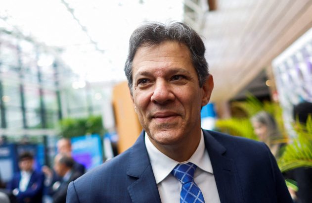 Homem de terno azul e gravata sorrindo e olhando para a direita em ambiente interno com fundo desfocado.