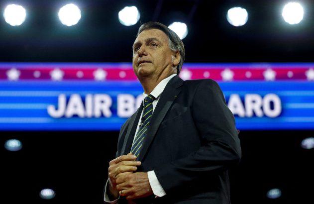 Jair Bolsonaro de terno, mãos juntas, em palco com seu nome e luzes ao fundo.