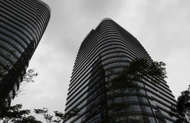 Dois arranha-céus de vidro escuro e curvo contra um céu cinzento. Árvores escuras na base.