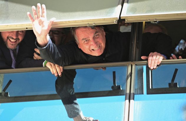 Homem sorridente acena de janela de veículo. Seu pé nu é refletido no vidro inferior. Outras pessoas.