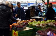Pessoas em mercado de rua comprando frutas frescas. Barracas exibem uvas e outras frutas em potes.