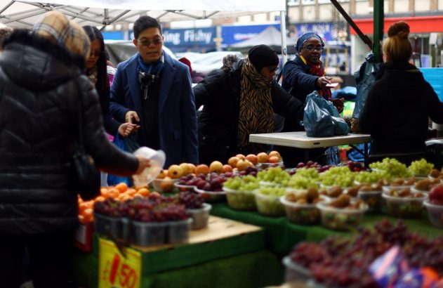 Pessoas em mercado de rua comprando frutas frescas. Barracas exibem uvas e outras frutas em potes.
