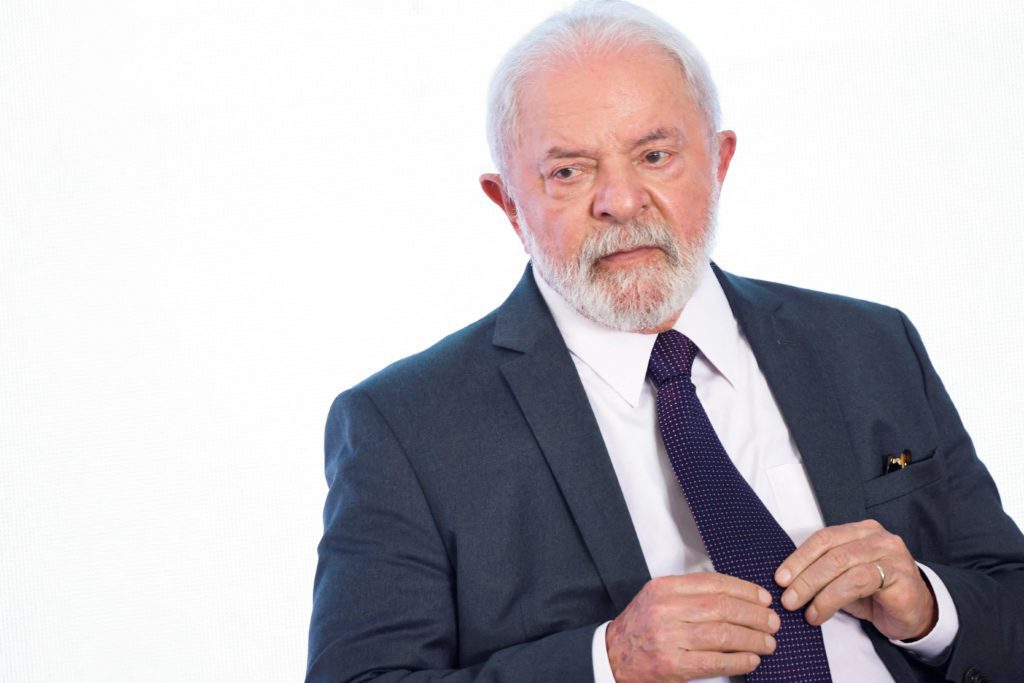 Homem de cabelos brancos e barba, de terno e gravata, ajustando a gravata. Fundo branco.