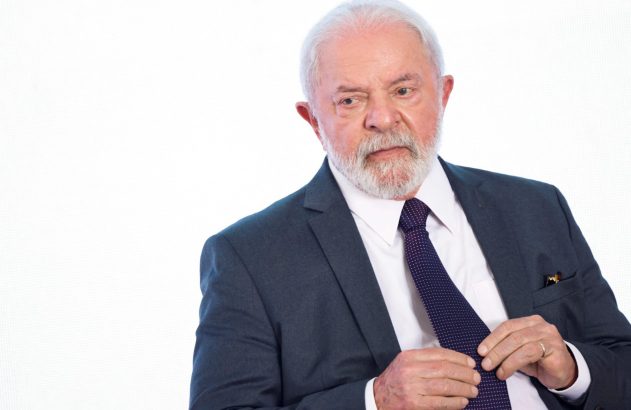 Homem de cabelos brancos e barba, de terno e gravata, ajustando a gravata. Fundo branco.