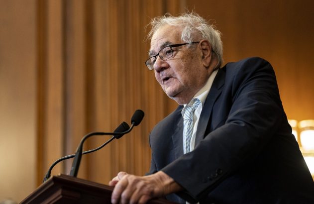 Homem sênior de óculos e terno discursa em pódio com microfones.