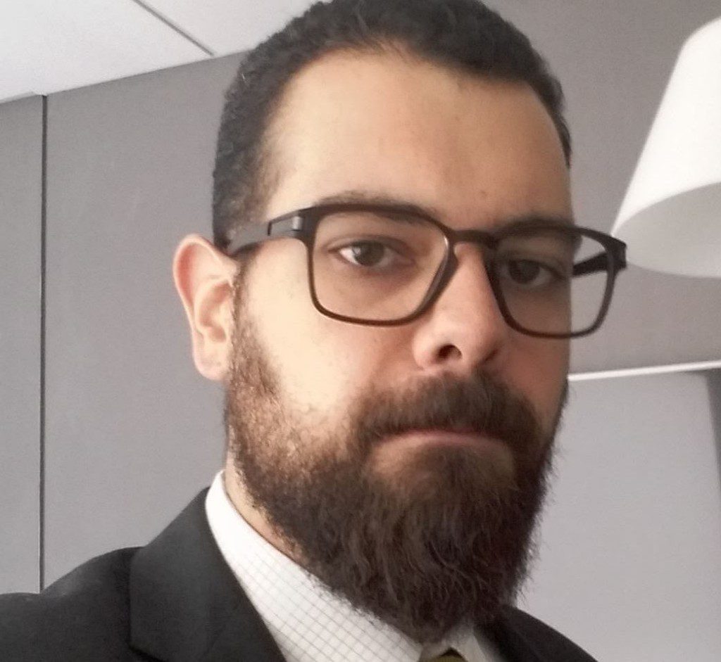 Homem barbudo de óculos e terno olhando para a câmera.