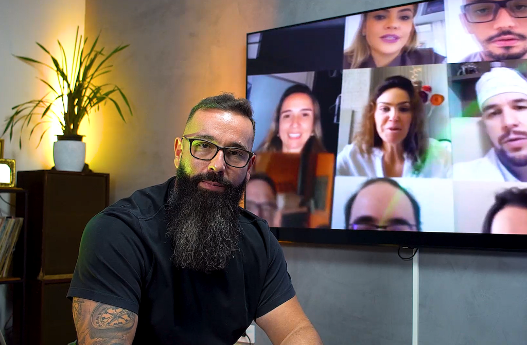 Homem barbudo e de óculos olhando para câmera. Ao fundo, TV exibe videoconferência com múltiplos participantes.