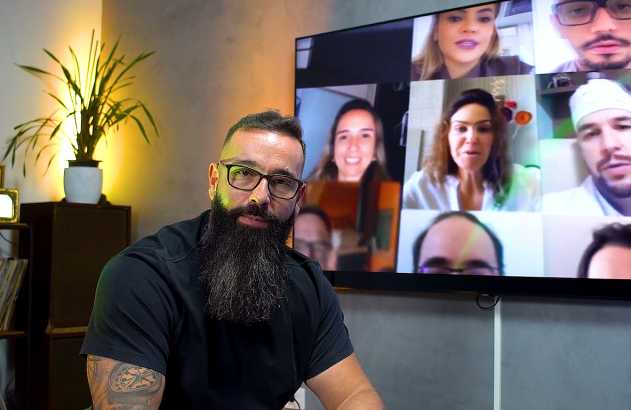 Homem barbudo e de óculos olhando para câmera. Ao fundo, TV exibe videoconferência com múltiplos participantes.