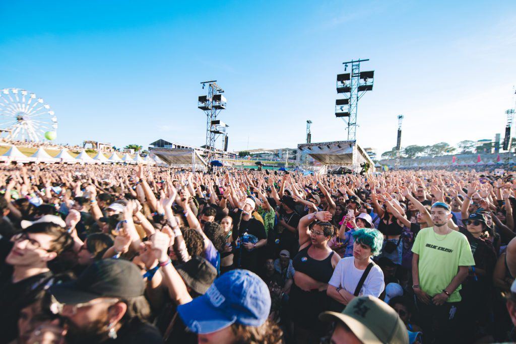 Multidão em concerto ao ar livre, mãos levantadas, palco e roda gigante ao fundo.